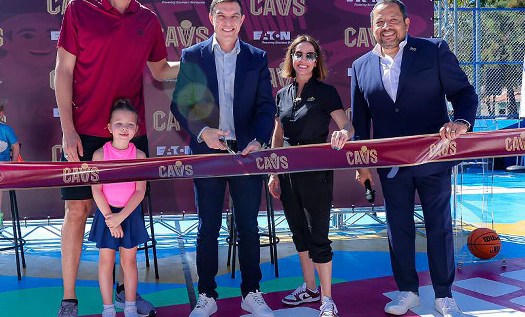 Four adults and a child stand behind a ceremonial ribbon on an outdoor basketball court during a Cleveland Cavaliers event, with a “CAVS” backdrop behind them.