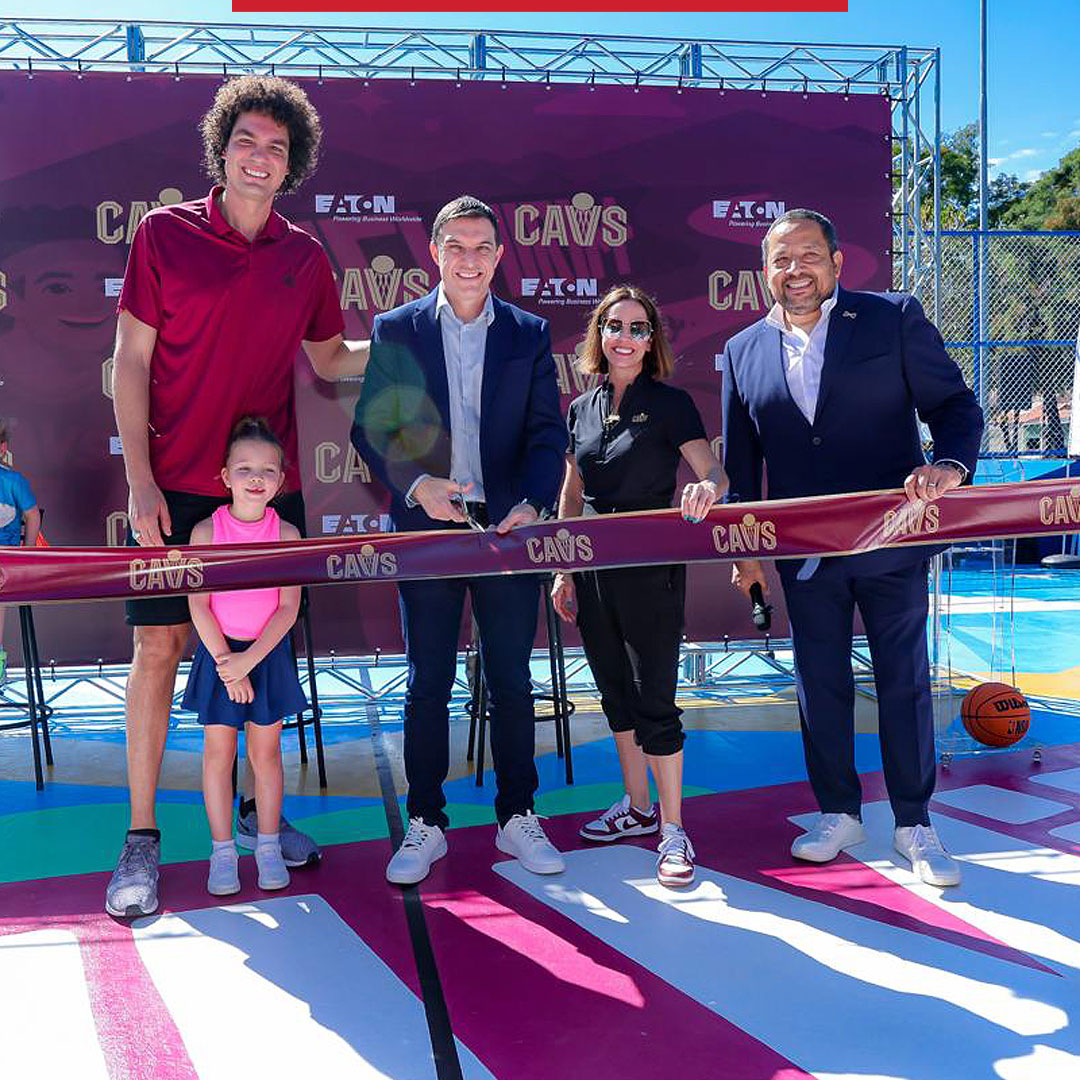 Four adults and a child stand behind a ceremonial ribbon on an outdoor basketball court during a Cleveland Cavaliers event, with a “CAVS” backdrop behind them.